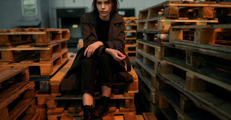 Pallet Projects - A Woman in Brown Coat Sitting on the Stack of Wooden Pallet while Looking at the Camera